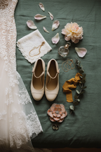 A bridal flat lay featuring a peony shaped blush pink ring holder, light beige vintage shoes and lace wedding gown on a green fabric surface with decorative elements like flowers, perfume bottle and ribbons.