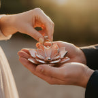 Hands placing an engagement ring into a handmade stained glass flower ring dish in blush pink tones with silver edges, captured in warm golden hour light; romantic proposal or wedding ceremony moment, ideal for ring bearer alternative, engagement photography, wedding styling, and meaningful gift for couple.