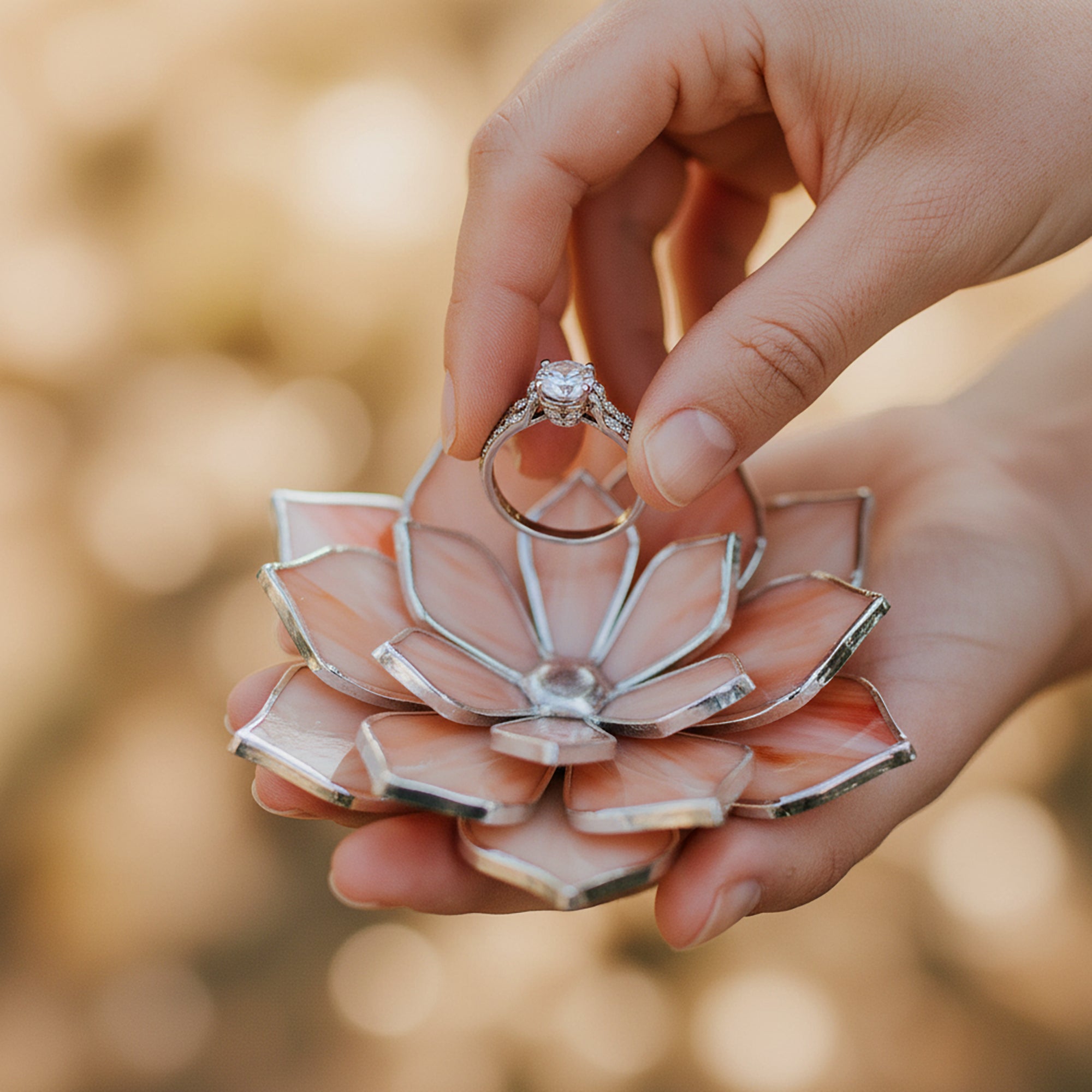 Two hands holding a lotus-shaped ring holder with a ring on it against a blurred background.