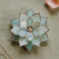 Handmade stained glass succulent ring dish in soft sage green and iridescent mint tones, silver-edged petals forming a layered floral shape with a central ring holder detail; decorative jewelry dish for rings, engagement flatlays, wedding styling, vanity decor, and meaningful gift for her, photographed in natural daylight on a neutral textured background.