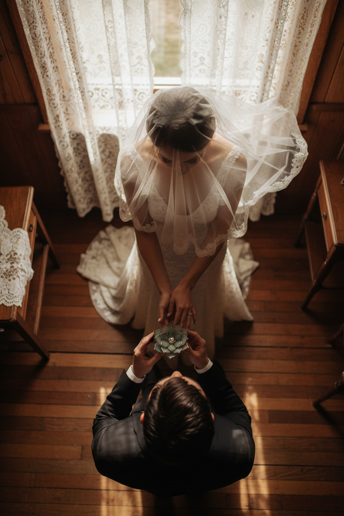 Woman in a white dress with a veil holding a flower shaped wedding ring bearer dish standing in front of a man on a wooden floor.