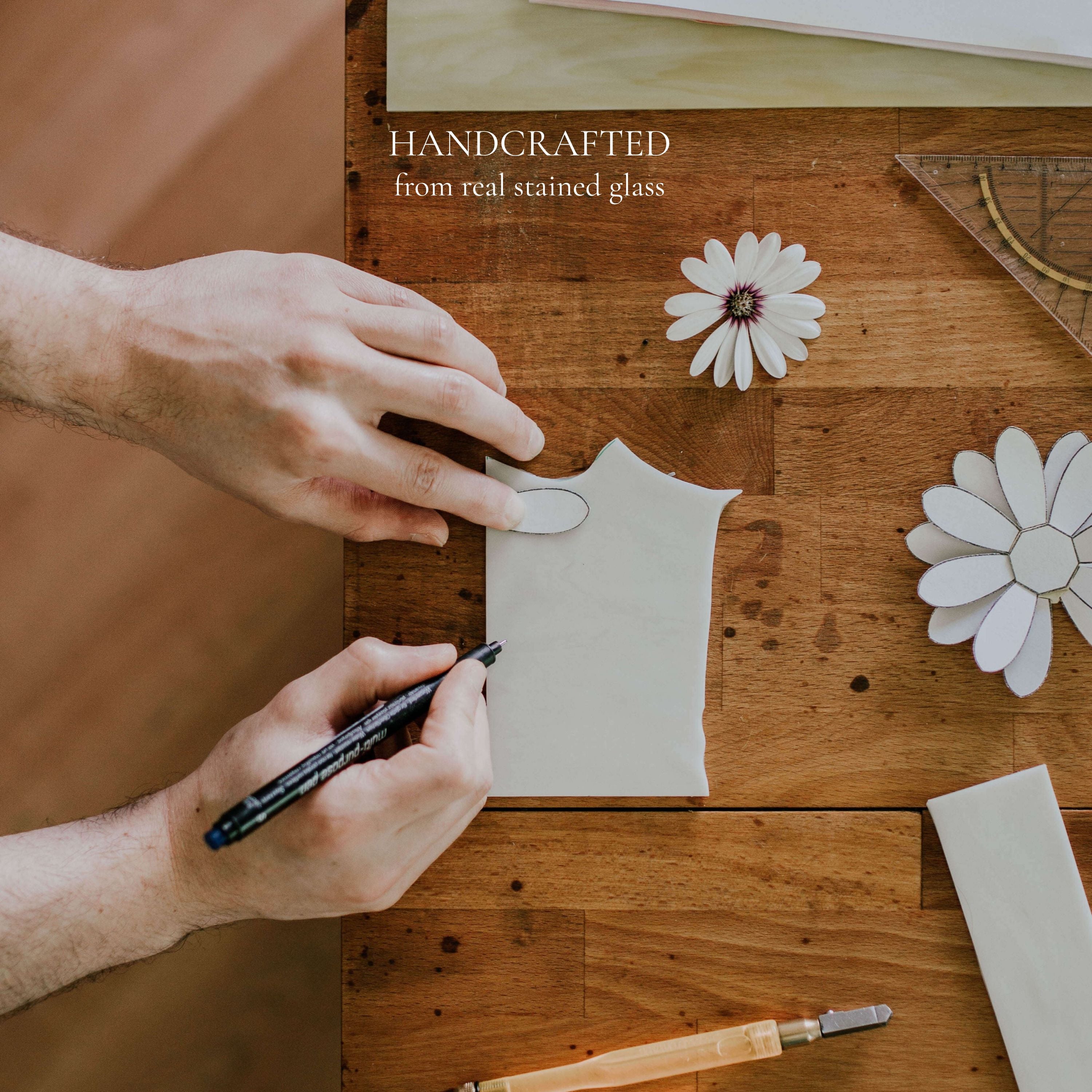 Artisan hand sketching a floral design for a handmade stained glass ring dish on a wooden workbench, highlighting real glass materials and handcrafted process; behind-the-scenes view of jewelry dish production, emphasizing craftsmanship, uniqueness, and quality for wedding ring holders and meaningful handmade gifts.