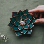 Hand placing an engagement ring into a handmade stained glass succulent ring dish in deep teal green with copper edges; floral jewelry holder with central ring slot, ideal for engagement ring display, wedding ring holder, ceremony styling, proposal photography, and unique gift for couple.