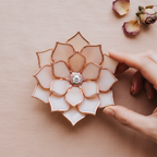 Hand touching a handmade stained glass flower ring dish in soft blush pink and ivory tones with copper edges, featuring a central engagement ring holder; minimal flat lay on neutral background, ideal for wedding ring display, engagement flat lay styling, proposal detail, and romantic gift for her or couple.