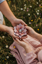 Two hands holding a pink flower-shaped glass wedding ring dish against a blurred natural background